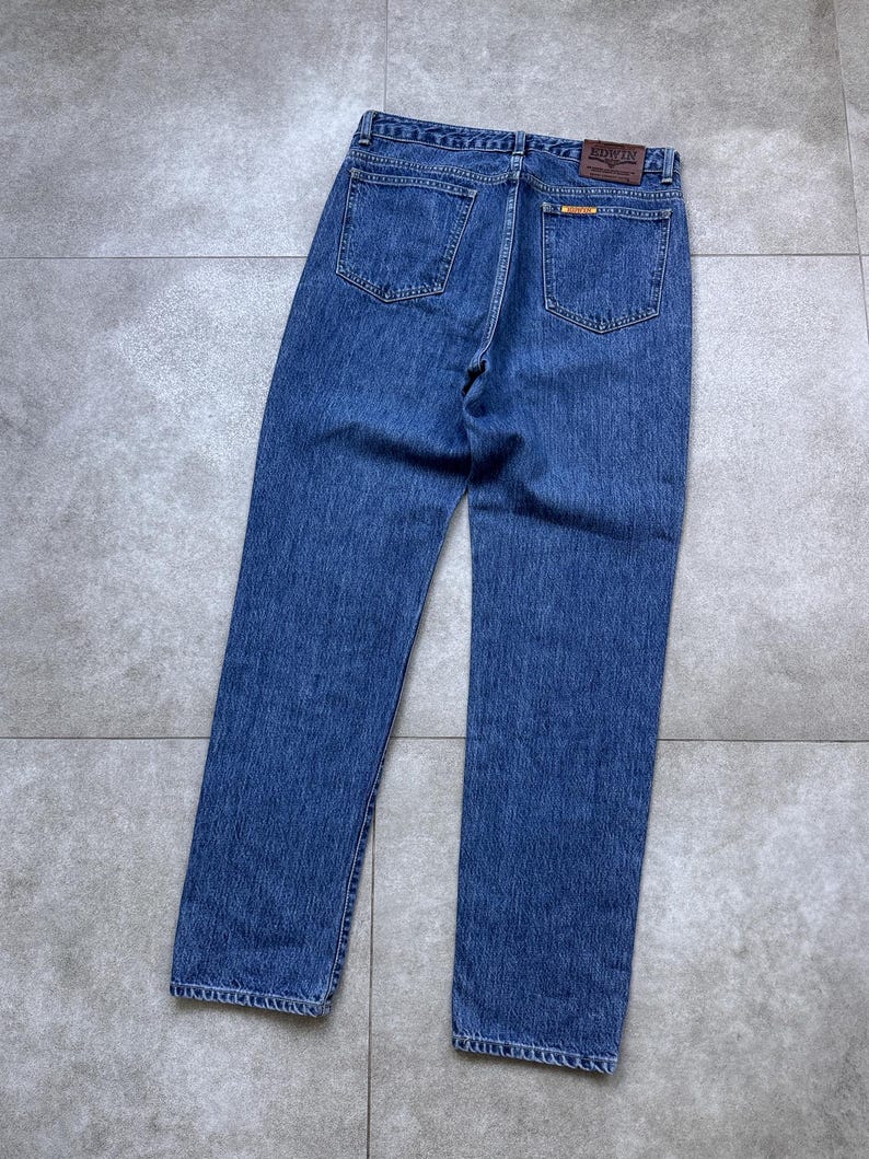 Pode incluir: Jeans azuis de ganga com um design cl&aacute;ssico de cinco bolsos. Os jeans apresentam um corte de perna reto e um patch de couro castanho com o nome da marca nas costas. Os jeans est&atilde;o estendidos numa superf&iacute;cie cinzenta.