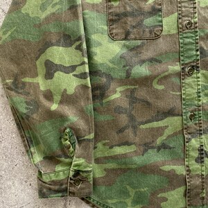 May include: A long-sleeved button-up shirt in a camouflage pattern with a green, brown, and black color scheme. The shirt has a patch pocket on the left chest and a button-up closure.