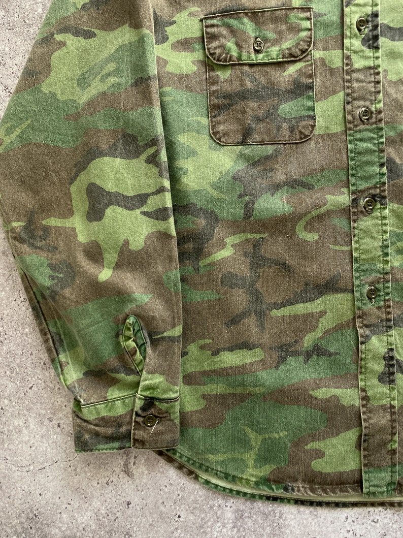 May include: A long-sleeved button-up shirt in a camouflage pattern with a green, brown, and black color scheme. The shirt has a patch pocket on the left chest and a button-up closure.