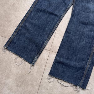 Puede incluir: Primer plano de unos vaqueros de mezclilla azul con corte recto. Los vaqueros tienen un dobladillo deshilachado y costuras visibles a lo largo de las costuras. La mezclilla tiene un aspecto descolorido, lo que sugiere desgaste.