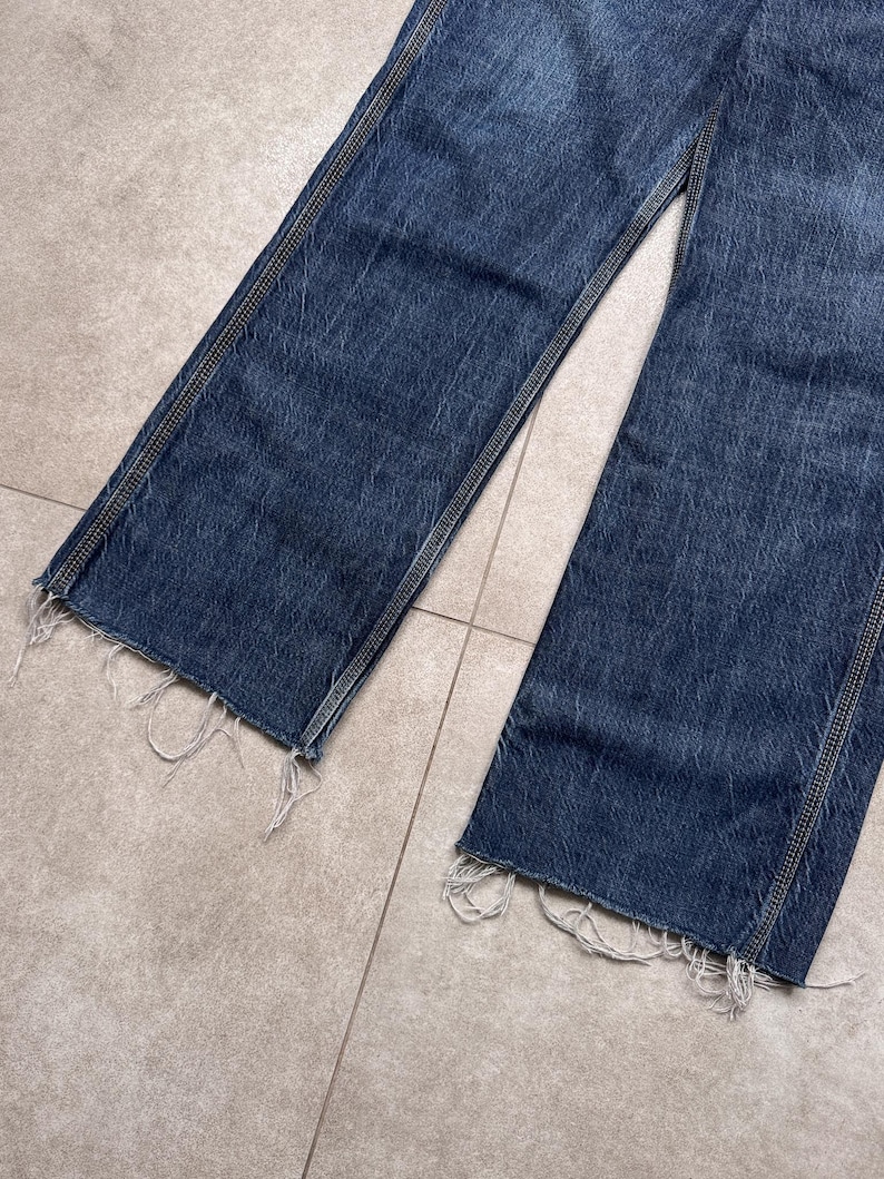 Puede incluir: Primer plano de unos vaqueros de mezclilla azul con corte recto. Los vaqueros tienen un dobladillo deshilachado y costuras visibles a lo largo de las costuras. La mezclilla tiene un aspecto descolorido, lo que sugiere desgaste.