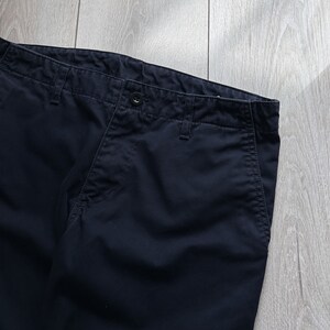 K&ouml;nnte beinhalten: Nahaufnahme einer marineblauen Hose. Die Hose hat einen Knopfverschluss und G&uuml;rtelschlaufen. Der Stoff scheint eine robuste Baumwolle oder Mischung zu sein. Die Hose ist gefaltet und zeigt Bund und Taschen. Der Hintergrund ist eine helle Holzmaserung.