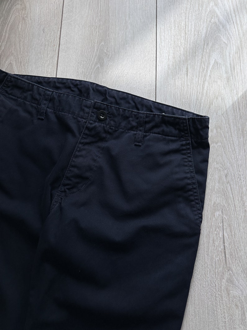 K&ouml;nnte beinhalten: Nahaufnahme einer marineblauen Hose. Die Hose hat einen Knopfverschluss und G&uuml;rtelschlaufen. Der Stoff scheint eine robuste Baumwolle oder Mischung zu sein. Die Hose ist gefaltet und zeigt Bund und Taschen. Der Hintergrund ist eine helle Holzmaserung.