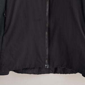 May include: A black jacket with a full-length zipper. The jacket has a textured fabric and a dark green patterned sleeve. The bottom hem of the jacket is slightly frayed.