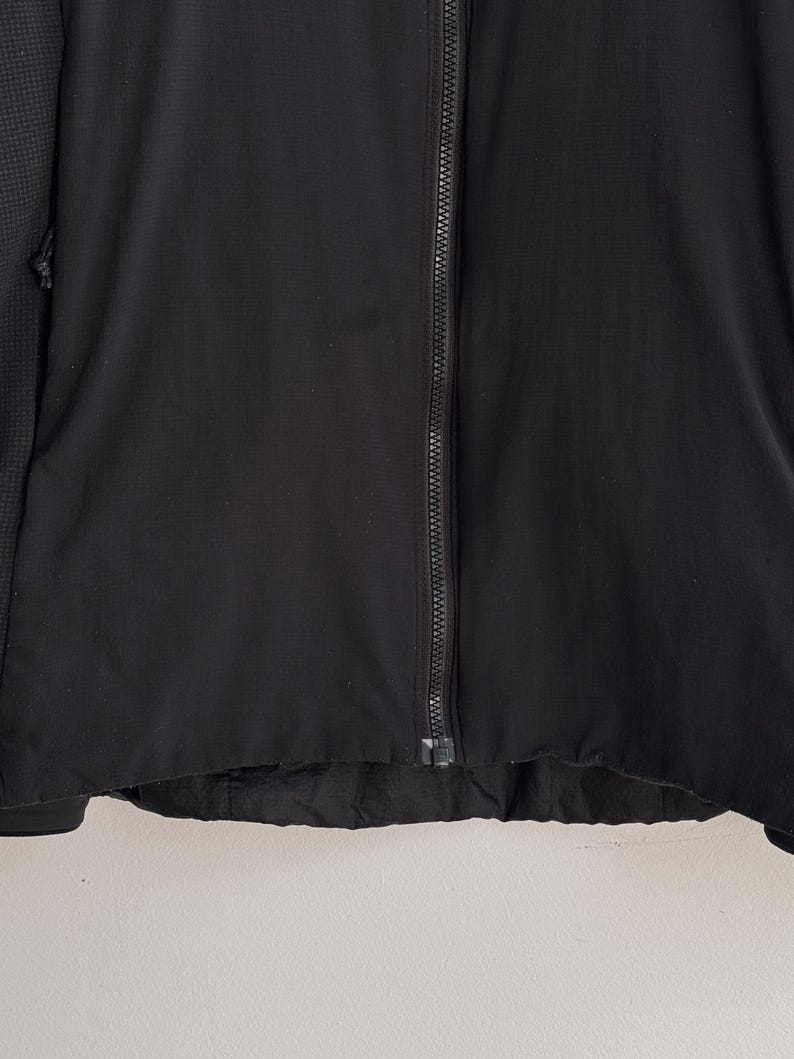 May include: A black jacket with a full-length zipper. The jacket has a textured fabric and a dark green patterned sleeve. The bottom hem of the jacket is slightly frayed.