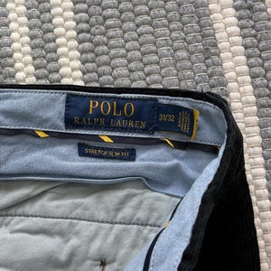 Puede incluir: Un pantal&oacute;n de pana negro Polo Ralph Lauren de ajuste delgado el&aacute;stico con una cintura a cuadros azul y blanco. La etiqueta dice "Polo Ralph Lauren" y "Stretch Slim Fit" con la talla "31/32".