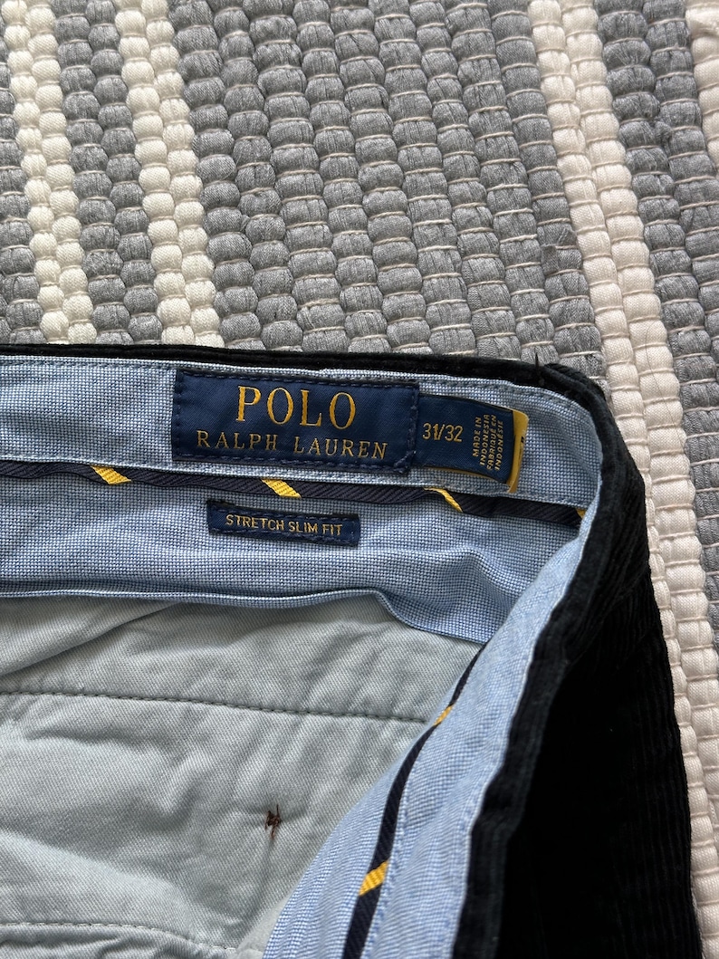 Puede incluir: Un pantal&oacute;n de pana negro Polo Ralph Lauren de ajuste delgado el&aacute;stico con una cintura a cuadros azul y blanco. La etiqueta dice "Polo Ralph Lauren" y "Stretch Slim Fit" con la talla "31/32".