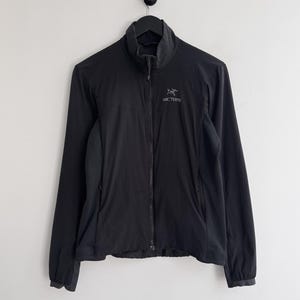 May include: Black full-zip jacket with a high collar. The jacket has long sleeves and a small logo on the chest. The jacket is hanging on a black hook against a white wall.