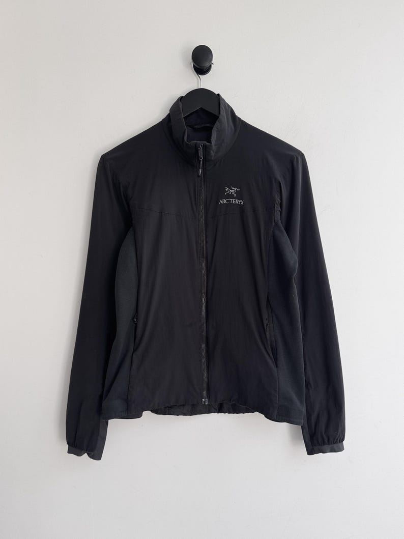May include: Black full-zip jacket with a high collar. The jacket has long sleeves and a small logo on the chest. The jacket is hanging on a black hook against a white wall.