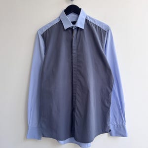 May include: A button-down shirt with a two-tone design. The shirt features light blue sleeves and a light blue collar, with a darker gray panel on the front. The shirt is hanging on a black hanger against a white wall.