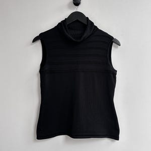 Puede incluir: Suéter negro sin mangas con cuello alto. El suéter presenta un cuello alto acanalado y detalles acanalados horizontales en el pecho. Cuelga de una percha negra sobre un fondo blanco.