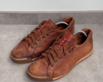 May include: A pair of brown leather trainers with matching brown laces and a low-top silhouette. The tongue features a red fabric tab with the white text PRADA. The side of the shoe is embossed with the brand name PRADA, and the soles are brown.
