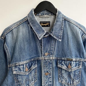 May include: A blue denim jacket with a Wrangler label. The jacket has a button closure, two chest pockets, and two front pockets. The jacket is distressed and faded.