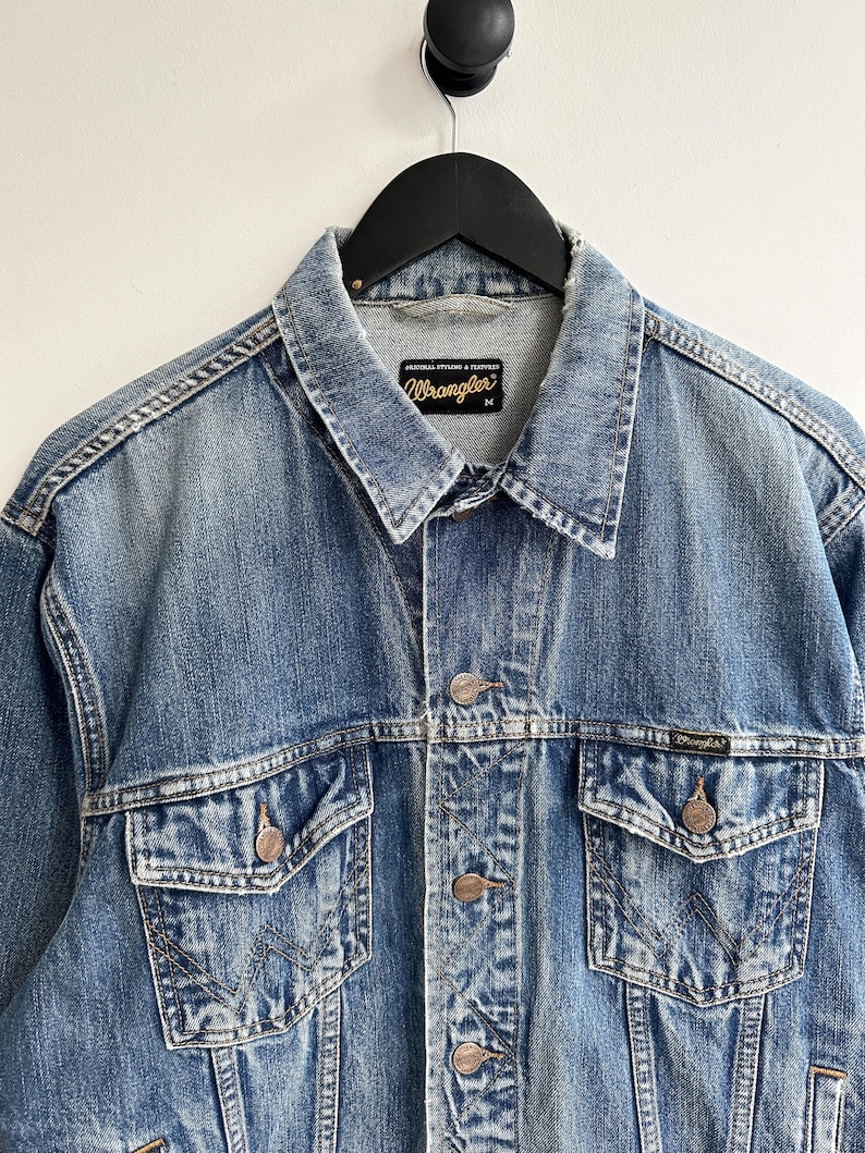 May include: A blue denim jacket with a Wrangler label. The jacket has a button closure, two chest pockets, and two front pockets. The jacket is distressed and faded.