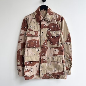 Pode incluir: Casaco militar camuflado bege e castanho, com quatro bolsos frontais e gola. O casaco está pendurado num cabide preto sobre um fundo branco. O padrão de camuflagem apresenta formas irregulares em vários tons.