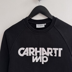 May include: Black crew neck sweatshirt with the white text "CARHARTT WIP" across the chest. The sweatshirt is hanging on a black hanger against a white wall. The Carhartt logo is on the neck.