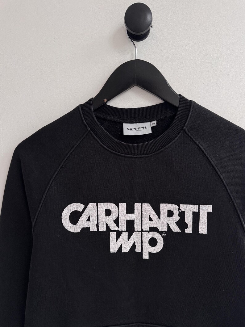May include: Black crew neck sweatshirt with the white text "CARHARTT WIP" across the chest. The sweatshirt is hanging on a black hanger against a white wall. The Carhartt logo is on the neck.
