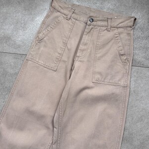 Puede incluir: Pantalones de color beige con grandes bolsillos delanteros y cierre de bot&oacute;n. Los pantalones tienen un dise&ntilde;o cl&aacute;sico e informal, adecuado para el uso diario. La tela parece ser de algod&oacute;n resistente o mezcla, con detalles de costura visibles.