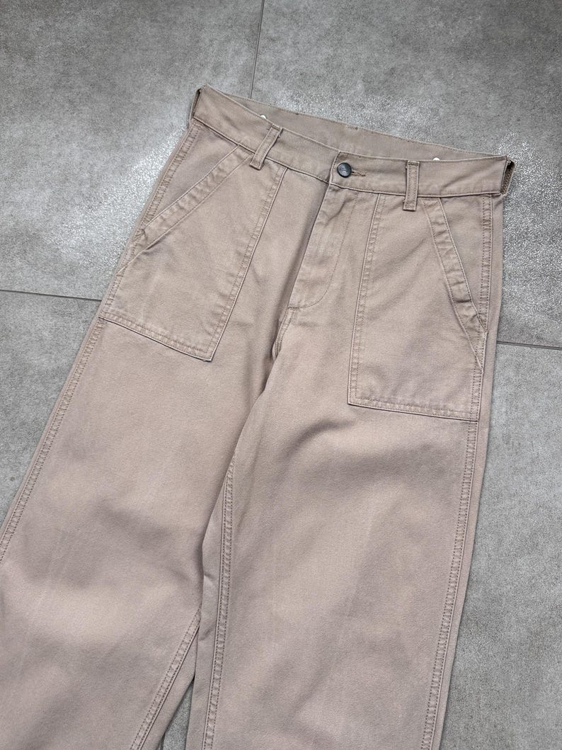 Puede incluir: Pantalones de color beige con grandes bolsillos delanteros y cierre de bot&oacute;n. Los pantalones tienen un dise&ntilde;o cl&aacute;sico e informal, adecuado para el uso diario. La tela parece ser de algod&oacute;n resistente o mezcla, con detalles de costura visibles.