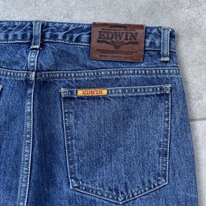 Pode incluir: Close-up de cal&ccedil;as de ganga azuis com um bolso traseiro e um remendo de couro castanho. O remendo apresenta a marca "EDWIN" e texto. Uma etiqueta amarela menor com "EDWIN" est&aacute; no bolso traseiro. As cal&ccedil;as de ganga s&atilde;o feitas de tecido de ganga dur&aacute;vel.