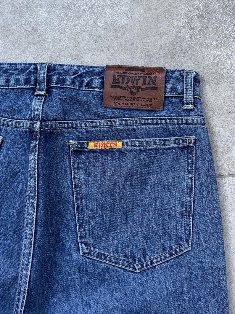Pode incluir: Close-up de cal&ccedil;as de ganga azuis com um bolso traseiro e um remendo de couro castanho. O remendo apresenta a marca "EDWIN" e texto. Uma etiqueta amarela menor com "EDWIN" est&aacute; no bolso traseiro. As cal&ccedil;as de ganga s&atilde;o feitas de tecido de ganga dur&aacute;vel.