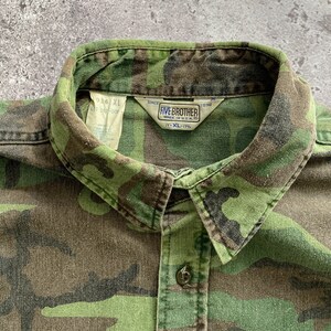 May include: A green, brown, and black camouflage button-up shirt with a Five Brother label. The label says "Since 1990" and "Made in U.S.A.". The size tag says "XL-17".