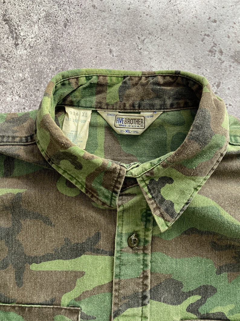 May include: A green, brown, and black camouflage button-up shirt with a Five Brother label. The label says "Since 1990" and "Made in U.S.A.". The size tag says "XL-17".