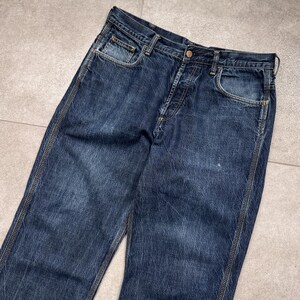 Puede incluir: Vaqueros de mezclilla azul oscuro con un dise&ntilde;o cl&aacute;sico de cinco bolsillos. Los vaqueros tienen cierre de bot&oacute;n y cremallera, trabillas para el cintur&oacute;n y costuras visibles a lo largo de las costuras. La mezclilla tiene un aspecto ligeramente descolorido.