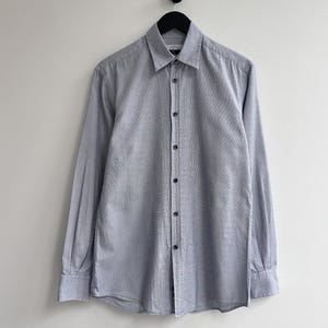 Könnte beinhalten: Hellblaues Langarmhemd mit dezentem Texturmuster, hängt an einem schwarzen Kleiderbügel. Das Hemd hat einen klassischen Kragen und ist mit dunkelblauen Knöpfen versehen.