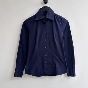 May include: A navy blue button-down shirt with long sleeves and a classic collar. The shirt is hanging on a black hanger against a plain white background. The shirt has a tailored fit and a smooth texture.