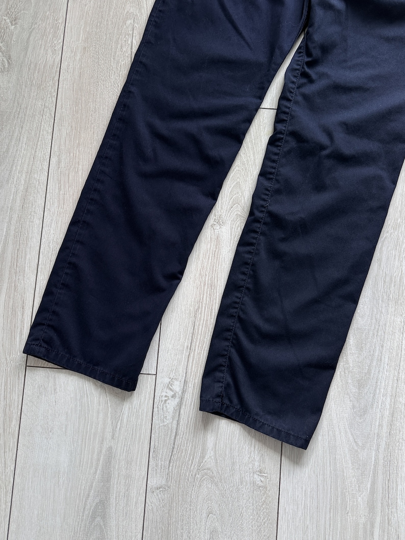 K&ouml;nnte beinhalten: Eine marineblaue Hose mit geradem Bein. Der dunkelblaue Stoff wird auf einer hellen Holzoptik-Oberfl&auml;che pr&auml;sentiert. Das Bild zeigt den unteren Teil der Hose, einschlie&szlig;lich S&auml;ume und N&auml;hte.