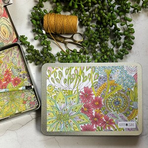 May include: A rectangular tin box with a floral design. The box is closed and has a white background with colourful flowers and leaves. The text "Kitty Jane" is printed on the box.