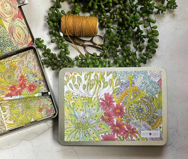May include: A rectangular tin box with a floral design. The box is closed and has a white background with colourful flowers and leaves. The text "Kitty Jane" is printed on the box.