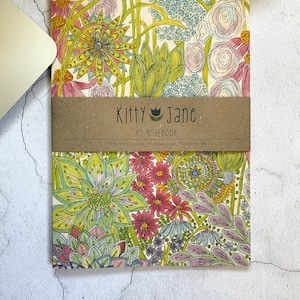 May include: A notebook with a floral design featuring a variety of flowers in shades of pink, yellow, green, and blue. The notebook is made of 100% recycled paper and has 38 unlined pages. The cover features the text "Kitty Jane AS NOTEBOOK".