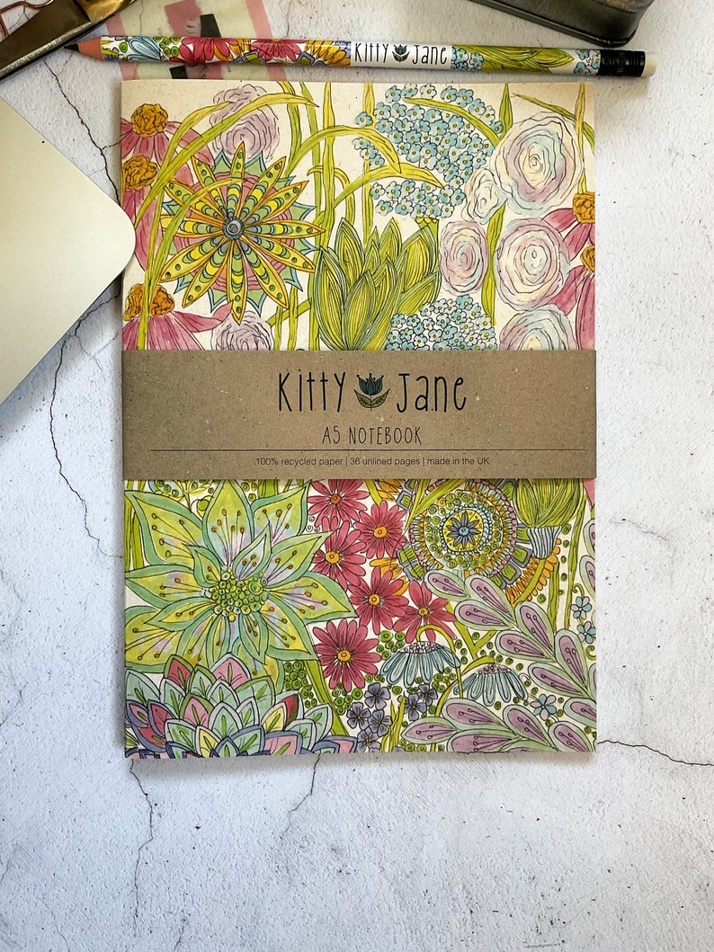 May include: A notebook with a floral design featuring a variety of flowers in shades of pink, yellow, green, and blue. The notebook is made of 100% recycled paper and has 38 unlined pages. The cover features the text "Kitty Jane AS NOTEBOOK".