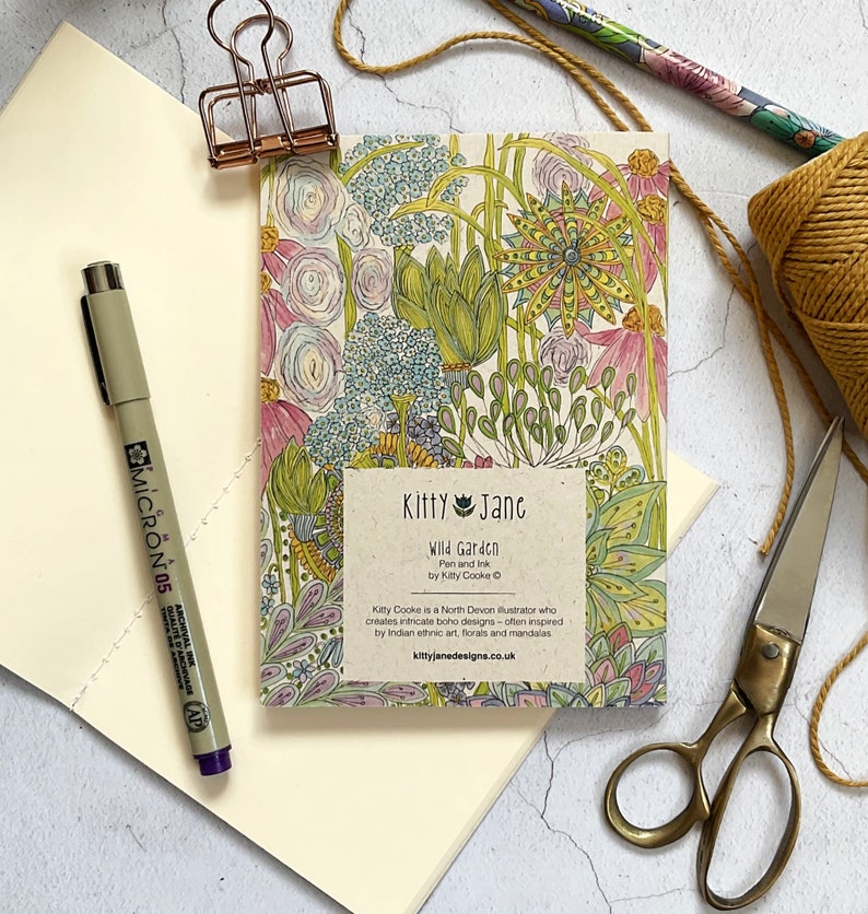 May include: A small notebook with a colourful floral design on the cover. The cover features a title that reads "Kitty Jane Wild Garden Pen and Ink by Kitty Cooke". The text below the title reads "Kitty Cooke is a North Devon illustrator who creates intricate boho designs - often inspired by Indian ethnic art, florals and mandalas kittyjanedesigns.co.uk".