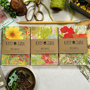 May include: Three small, colourful floral notebooks with brown paper covers that say "Kitty Jane Mini Notes" and "50 blank pages | FSC paper | made in the UK".