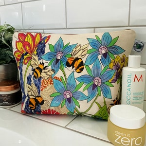 May include: A white canvas makeup bag with a floral print featuring blue flowers, green leaves, and yellow bees. The bag has a zipper closure.