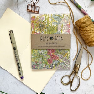 May include: An A6 notebook with a floral design featuring pink, green and blue colours. The notebook cover has the text "Kitty Jane A6 Notebook 100% recycled paper | 36 unlined pages | made in the UK."
