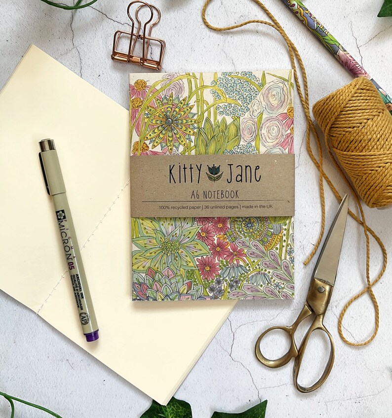 May include: An A6 notebook with a floral design featuring pink, green and blue colours. The notebook cover has the text "Kitty Jane A6 Notebook 100% recycled paper | 36 unlined pages | made in the UK."