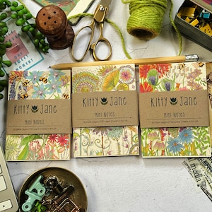 May include: Three small floral patterned notebooks with brown paper covers. Each notebook has the text "Kitty Jane Mini Notes" and "50 blank pages | FSC paper | made in the UK".