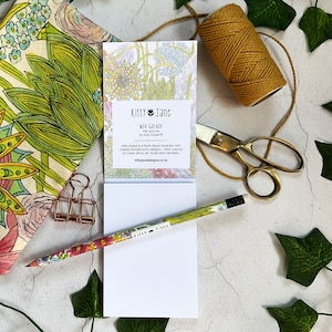 May include: A white notepad with a floral design on the front. The notepad has the text "Kitty Jane Wild Garden Pen and Ink by Kitty Cooke" and a website address. The notepad is surrounded by green leaves, a gold pencil, a gold paperclip, and a spool of twine.