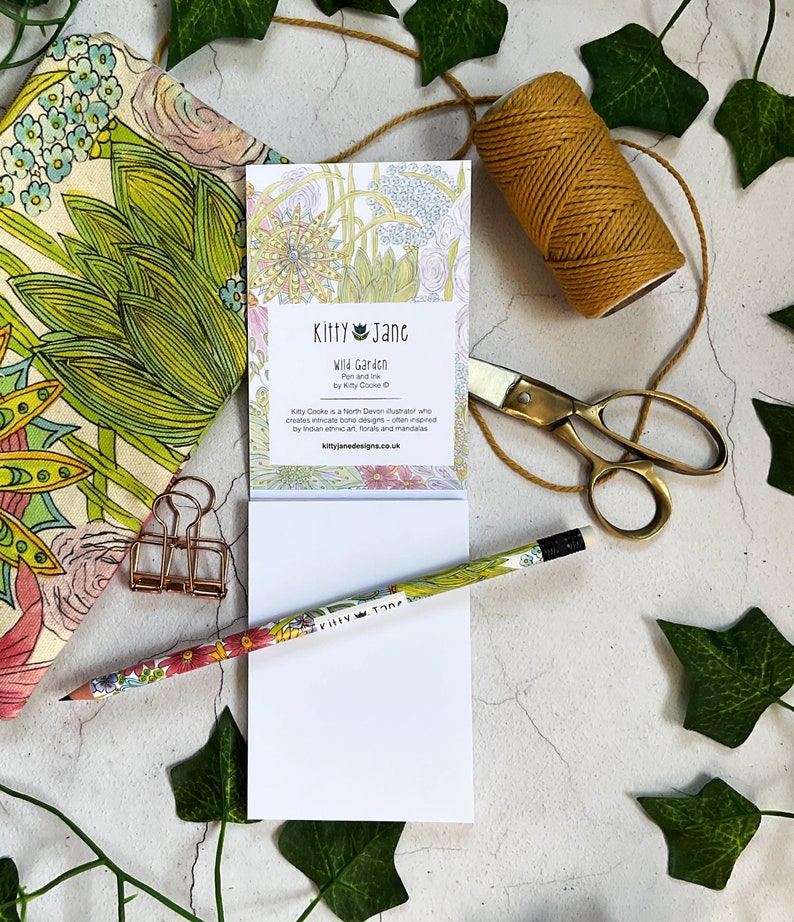 May include: A white notepad with a floral design on the front. The notepad has the text "Kitty Jane Wild Garden Pen and Ink by Kitty Cooke" and a website address. The notepad is surrounded by green leaves, a gold pencil, a gold paperclip, and a spool of twine.