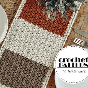 May include: A crocheted table runner with three stripes in brown, cream, and orange. The runner is on a wooden table with two white plates and silverware. The text "crochet pattern" and "The Turtle Trunk" is on a white circle.