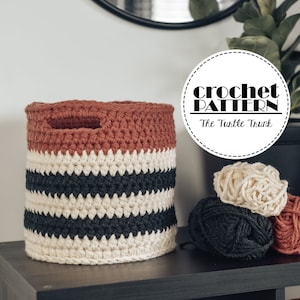 May include: A crocheted storage basket with a striped pattern in black, white, and rust orange. The basket has a handle and is sitting on a black shelf. There are three balls of yarn in the colors black, white, and rust orange. The image also includes a round sticker with the text "crochet pattern" and "The Turtle Trunk".