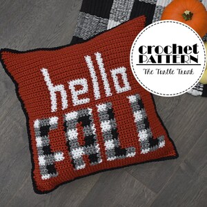 May include: A red crochet pillow with the words "hello fall" in white and black checkered letters. The pillow is on a wood floor with a black and white checkered fabric and two pumpkins in the background.