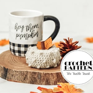 Op de afbeelding: Haakpatroon voor een kleine, beige pompoen met een bruine steel en oranje bladeren. De pompoen ligt op een houten schijf met een mok waarop "hey there pumpkin" staat en een dennenappel.