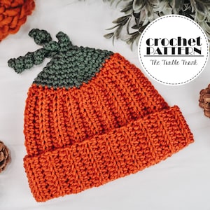 A ribbed, crochet Pumpkin Beanie laying on a white surface with a scarf, pine cones, and plant around it.