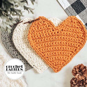 May include: Three crocheted heart-shaped coasters in gray, white, and orange. The coasters are arranged in a row, with the orange coaster in the front. The text "crochet pattern" and "The Turtle Trunk" is visible in a circle.