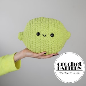 May include: A crocheted lime-shaped plush toy with a smiling face. The toy is green and has black eyes and a mouth. The toy is being held in a hand.